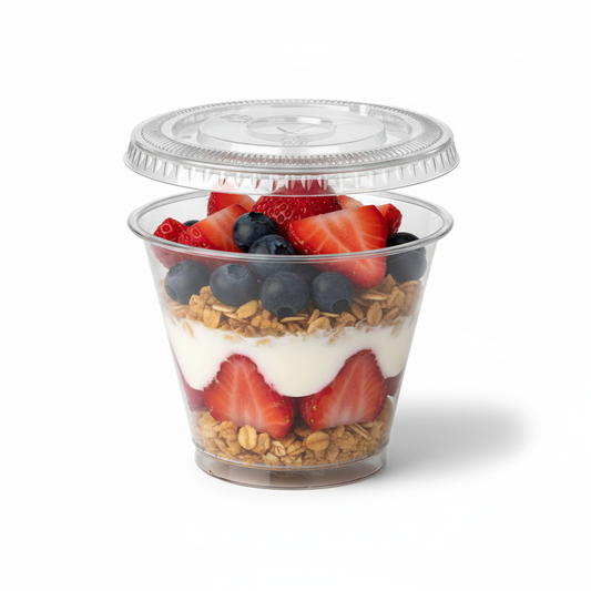 Le couvercle plat transparent en plastique PET de 90 mm est montré clairement. Il est posé sur un pot à dessert 25 cl, lui aussi transparent et rempli d'un dessert coloré (par exemple, une mousse ou une salade de fruits), illustrant la fonction de protection et l'herméticité. Le fond est blanc épuré, avec un éclairage qui accentue la transparence et la brillance du couvercle.