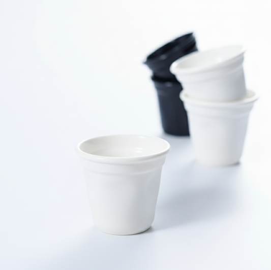 Photographie studio d'une verrine blanche au premier plan, avec une mise au point nette. En arrière-plan flou, des piles de verrines noires et blanches ajoutent de la profondeur.